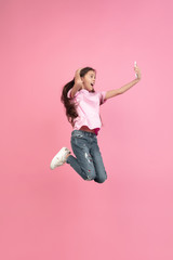 Jumping high, selfie. Caucasian little girl portrait isolated on pink studio background. Cute brunette model in shirt. Concept of human emotions, facial expression, sales, ad, childhood. Copyspace.