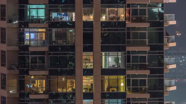 Rows of glowing windows with people in the interior of apartment building at night. Modern skyscraper with glass surface. Concept for business and modern life. Pan right
