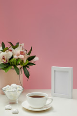 Fototapeta na wymiar photo frame, orchid flowers, cup of tea and sweets on the pink background. empty space for text. mock up with copy space.