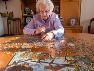 An elderly lady sits at a table placing a jigsaw piece into a puzzle.She has a gin and tonic in one...