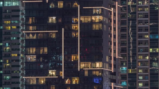 Rows of glowing windows with people in the interior of apartment buildings at night. Modern skyscrapers with glass surface. Concept for business and modern life. Zoom out