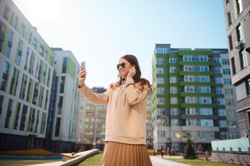 Young stylish smiling long-haired brunette uses a cellphone and headphones on the street. Woman...