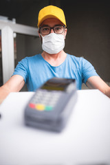 Fototapeta na wymiar Deliveryman with protective medical mask holding pizza box and POS wireless terminal for card paying - days of viruses and pandemic, food delivery to your home and safety hygiene measures.