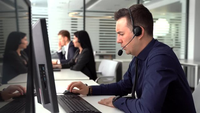 smiling man taking call in busy call center. In the background there are his colleagues also talking to clients on phone