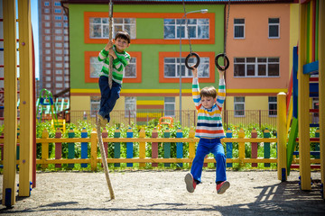 Two brothers sibling boy in casual clothes on playground climbing rope and athletic rings. Kids has...