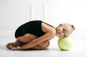 Young girl athlete rhythmic gymnastic in a black suit lyong on the floor, head on the ball, looking at camera smiling - obrazy, fototapety, plakaty