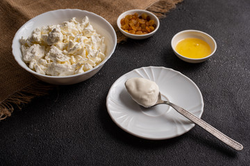 Cottage cheese in a white plate with a spoon of sour cream on a dark background. Farm products, close-up.