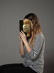 Happy world book day 2020, be safe and read to become someone else - woman covering face with book in mask while reading on grey studio background. Celebrating, education, art, protection concept.