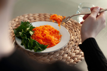 Caucasian woman eating green healthy tasty eco salad on dining table. Wicker mat and glass of water on table. Parsley and carrots on plate.