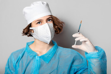 A young nurse in a protective mask, a blue coat and white latex gloves holds a syringe. Gray...