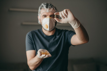 brunette white european man doctor with medical mask on gray background holding a pill of vitamin omega 3. Coronavirus, illness, infection, quarantine, surgical bandage, respirator with valve.