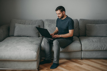 Young white european brunette man sitting on gray couch at home with black laptop, working in freelance, distant earnings.