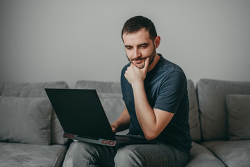 Young white european brunette man sitting on gray couch at home with black laptop, working in freelance, distant earnings.