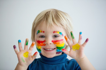 Beautiful blond toddler boy with rainbow painted on his face and messy hands