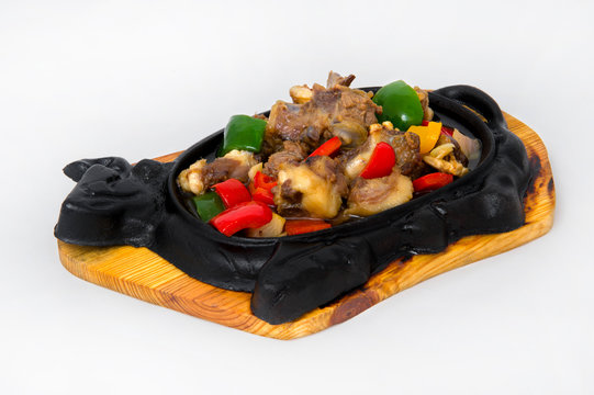 Fatty pieces of beef stew with separately fried colored sweet pepper mixed in a cast iron plate in the shape of a cow on a wooden stand on a white background