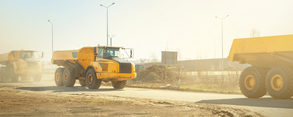 Many big articulated heavy industrial yellow dumper trucks driving on new highway road construction site on sunny day with blue sky background. Construction equipment machinery working on open pit - obrazy, fototapety, plakaty