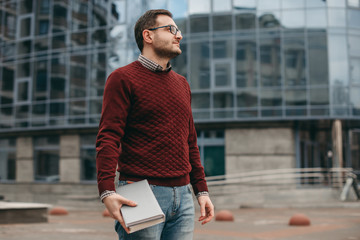 Portrait of a man in glasses for vision wearing a shirt and sweater, with , frame, with books, notebook in his hands, on the background of a modern building. Education, institute, business, outside