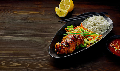 Asian- style chicken with savory and sweet teriyaki sauce, rice, sesame, salad, edamame, lemon and sweet chili sauce on a wooden table.