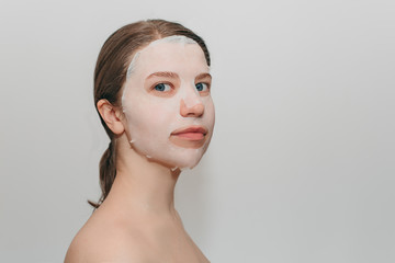 Portrait of young beautiful european woman with blue eyes with collagen silicone fabric with white mask on face on white background. Beauty treatments, the secret of youth, beauty, health.