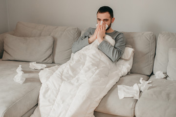 Young sick bearded european man sneezes into napkin at home on gray sofa with white blanket. Disease, protection, coronavirus, virus, disease, flu, respiratory dressing.