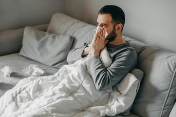 Young sick bearded european man sneezes into napkin at home on gray sofa with white blanket....