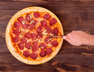 Salami pizza with mushrooms, on bamboo bottom, on wooden table hand cutting pizza, top view