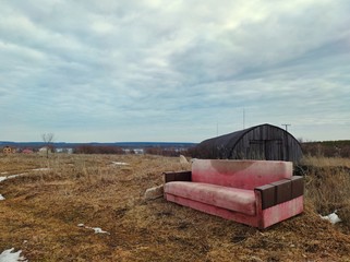 a lonely old sofa in a field against the background of an abandoned hangar and a blue sky with...