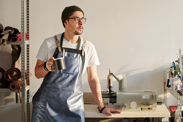 Young restful craftsman in leather apron and beanie hat holding mug with drink