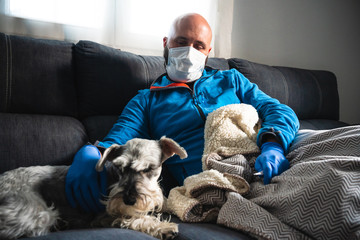 Sick man with procedure mask and medical gloves checking a thermometer liying on the sofa and caress a dog