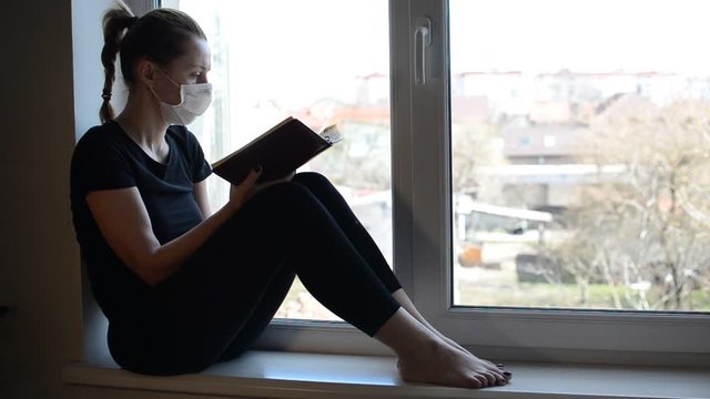 A young girl in a medical mask sits on a windowsill near a window and reads a book