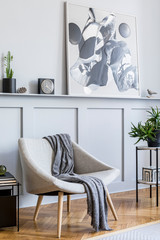 Stylish interior of living room with design grey armchair, plaid, marble stool, paintings, cacti, plants, decoration, black clock and elegant personal accessories in modern home decor.