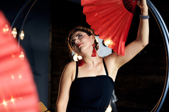 Young brunette woman, wearing little black dress, holding red paper fans in both hands, doing flamenco posing in dark photo studio with lamps and lights. Passionate female dancer.