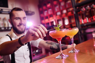the bartender holding two glasses with an orange alcoholic cocktail smiles against the night club...