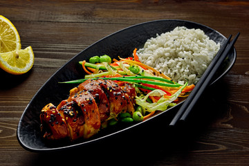Delicious Asian teriyaki chicken, salad made of cucumber, carrot, ginger, pepper and cabbage, edamame, sesame and rice in a black pottery plate.