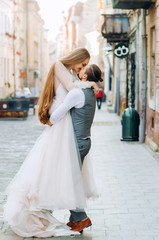 Amazing groom holds his bride in hands kissing on lips against morning street background