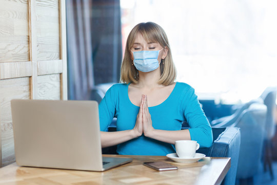 Portrait of calm young blonde woman with surgical medical mask in blue t-shirt is sitting and working on laptop and try to relax in yoga pose. Indoor working, medicine and health care concept.