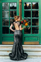 Pretty girl in black evening dress with open back holds a bouquet in her hand on green door...