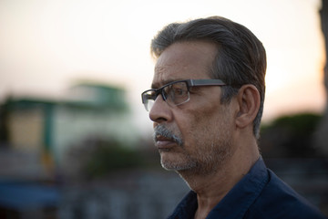 Naklejka premium Portrait of an An old/aged Indian Bengali man in blue shirt is standing on a rooftop under the open sky in front of a urban landscape. Indian lifestyle