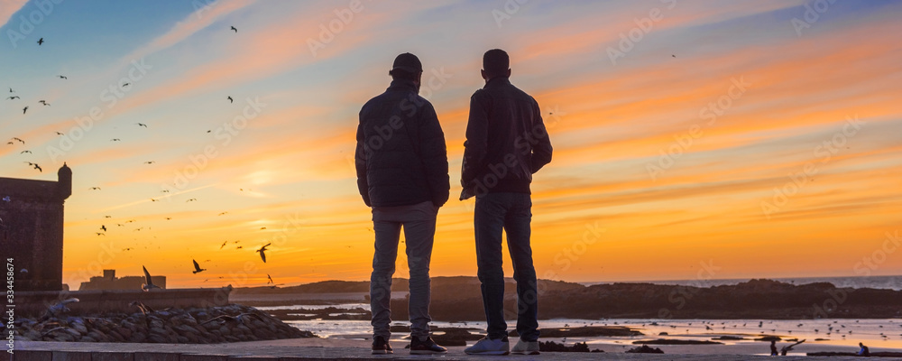 Wall mural panorama view of the silhouette from two people at the old fort essaouira on sunset, blue sky, backg