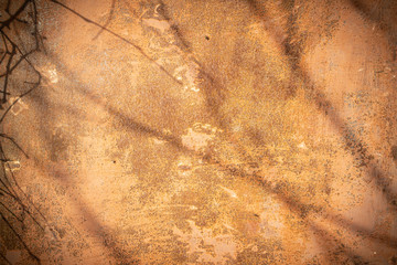 Textural old metal orange background in grunge style, covered with peeling paint and rust. Illuminated by the sun with shadows of branches and a faint dark vignette.