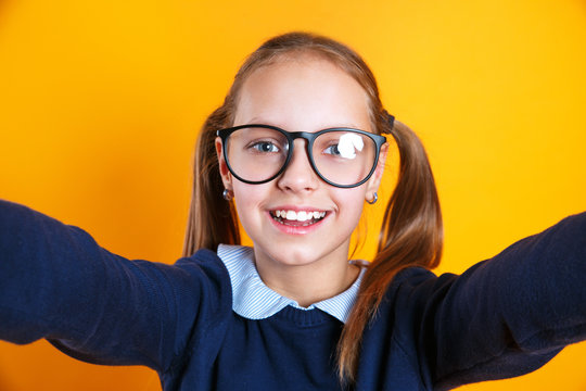 Close up of little kid girl 12-13 years old in glasses doing selfie on mobile phone on yellow background.