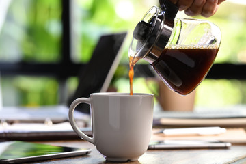 A hand pouring steaming coffee in to a cup on a work desk when work from home - 334462257