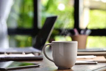 A hot and steaming cup of coffee on a desk with laptop and gadget when work from home