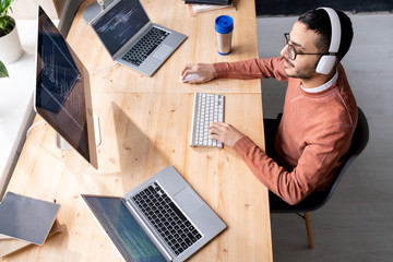 Busy young software developer in headphones sitting by table and typing