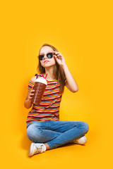 Cute caucasian young girl in t-shirt and sunglasses drinking milk cocktail on yellow background