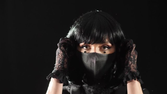 A girl in an evening look with black hair and black lace gloves, removes a medical mask from her face under which a black elegant mask of sequins and rhinestones is depicted