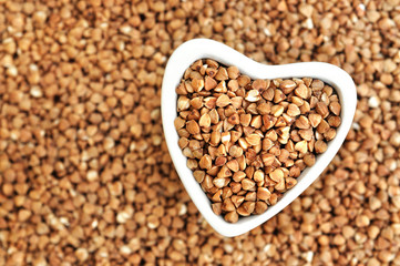 Heart-shaped bowl of roasted buckwheat on groat background, gluten free ancient grain for healthy diet