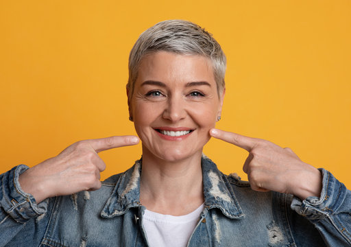 Perfect Smile. Mature woman pointing on strong healthy teeth, yellow background