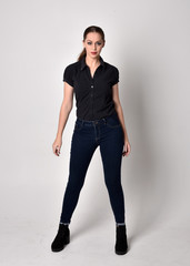 Portrait of a pretty brunette girl with ponytail, wearing jeans boots and a black blouse.  full length standing pose on a studio background.