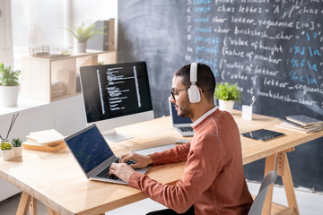 Busy young programmer with headphones analyzing data on laptop display